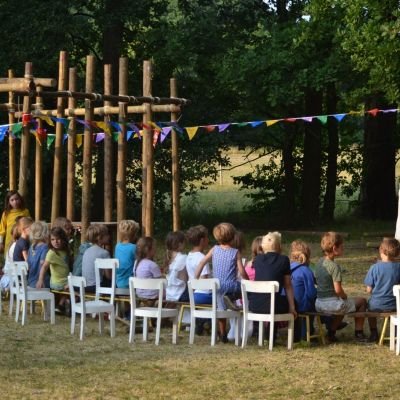 kapoenen op stoeltjes op een grasveld. Naast de kinderen staat een sjorconstructie versiert met vlaggetjes.