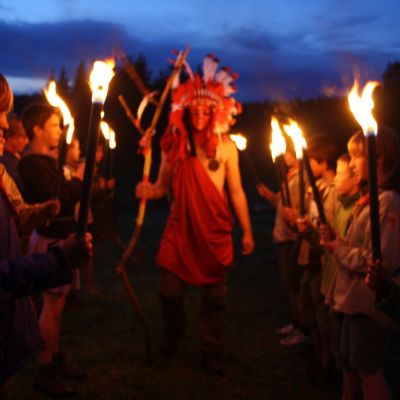 Bij schemering staat scouts in twee rijen, gezicht naar elkaar met fakkel in de hand. tussen de twee rijen loopt een gevederde indiaan.