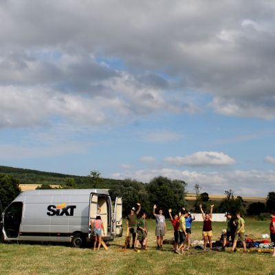 Givers staan klaar om een grijze camionette uit te laden. Ze zwaaien naar de camera