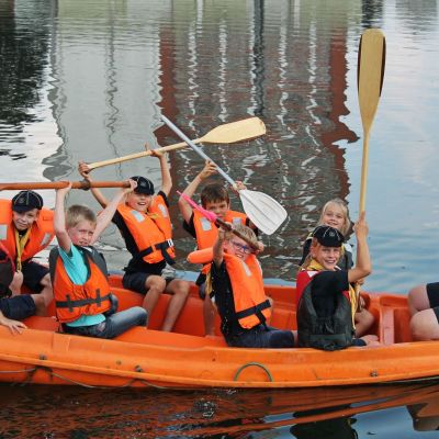 acht zeewelpen in een oranje bootje. Op de boeg zit een leider. Ze zwaaien met hun roeispanen.