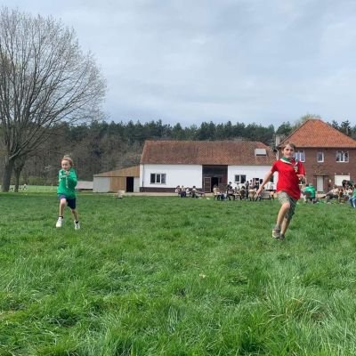 Leden lopen op een scoutsterrein, op de achtergrond zie je het lokaal