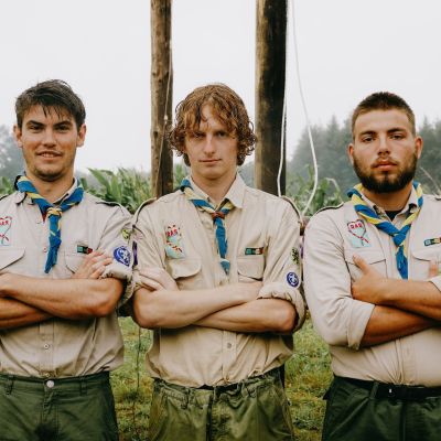 drie scoutsleiders staan op een rijtje, armen over elkaar, op kamp
