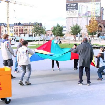 Leiding en kinderen uit de buurt spelen op een pleintje. Op de voorgrond staat een grote Uitwijker-koffer met spelmateriaal in.