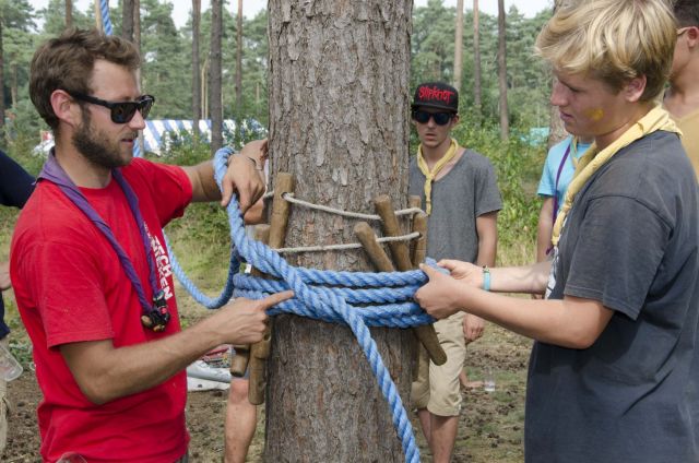 Twee scoutsleiders steken een paar takken tussen de bast van een boom en het touw.