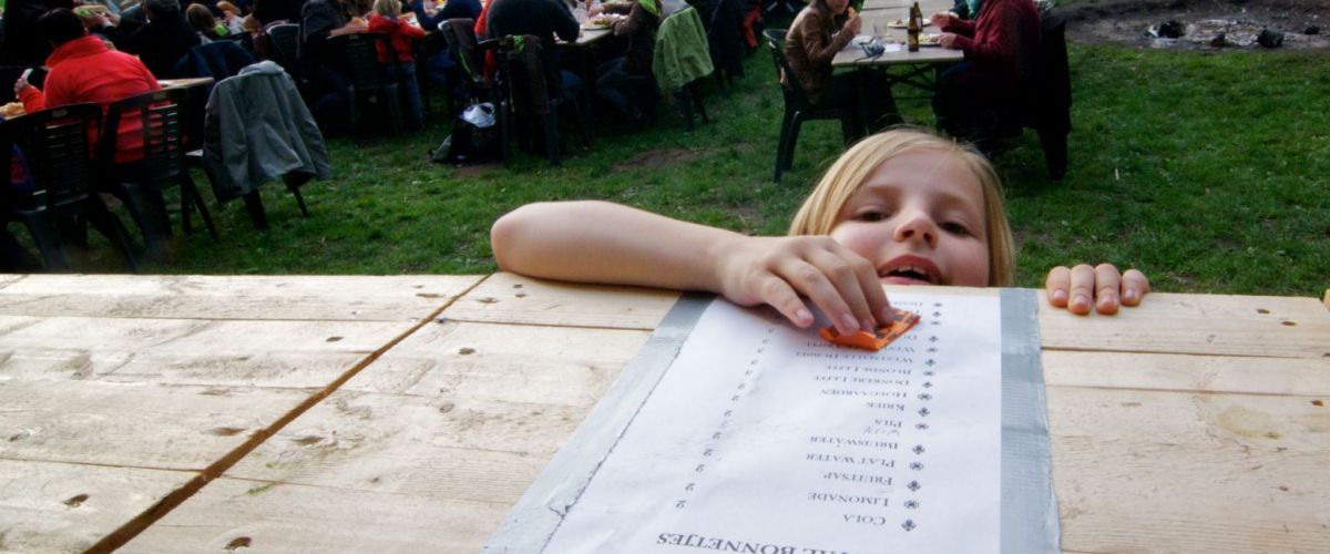 Op de achtergrond zitten mensen aan lange tafels, vooraan komt een kindje bestellen aan de toog, hoofd net boven de tafel.