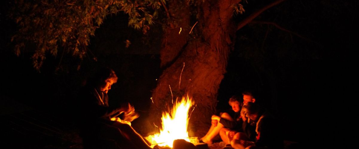 Scoutsleider leest voor aan drie leden rond het kampvuur