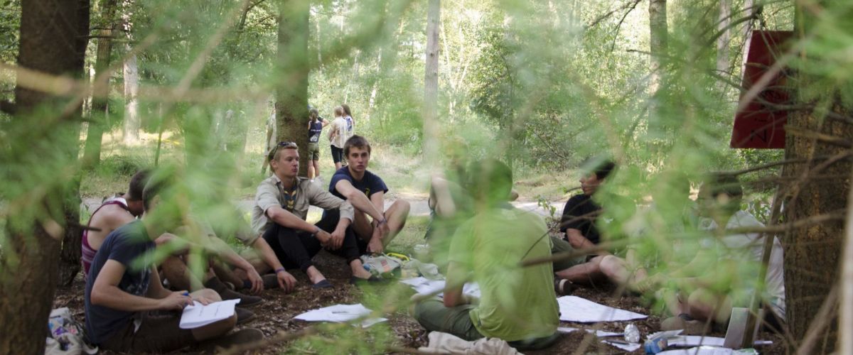 Sabine De Schutter scouts zitten op de bosgrond, ze lijken een heuse vergadering te houden in het bos.