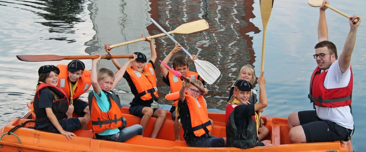 acht zeewelpen in een oranje bootje. Op de boeg zit een leider. Ze zwaaien met hun roeispanen.