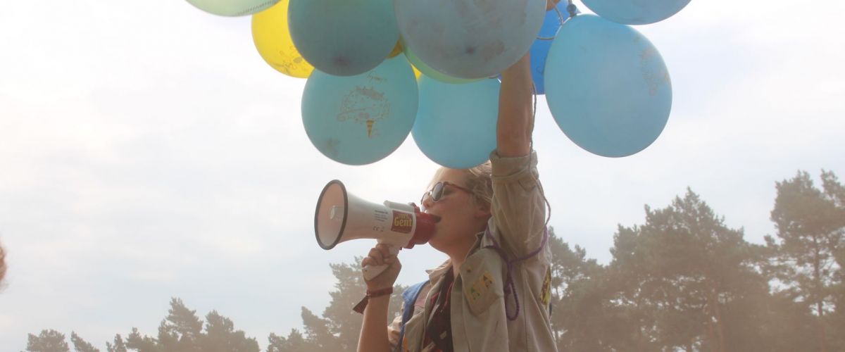 Meisje met megafoon en tros ballonnen zit op schouders van jongen.