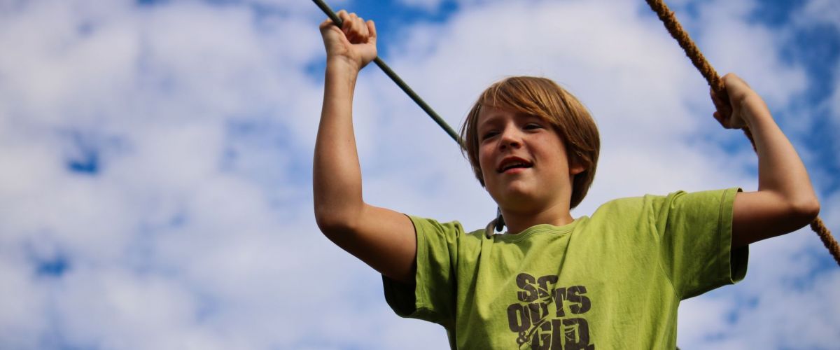 Een scout met groene scoutsshirt balancerend op een touwbrug