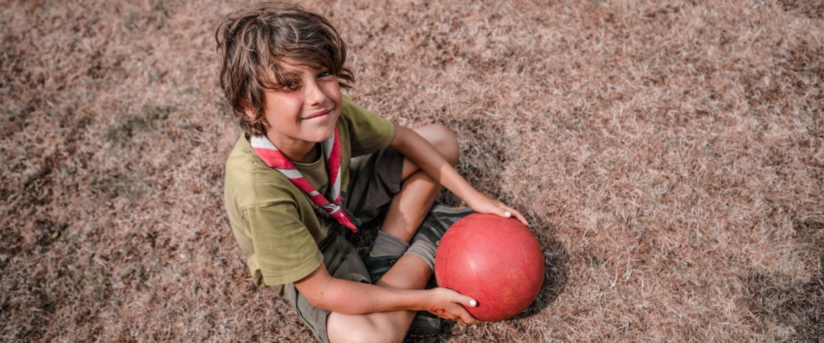 jongen met groene scoutst-shirt en korte groene broek zit in het gras met een rode bal.