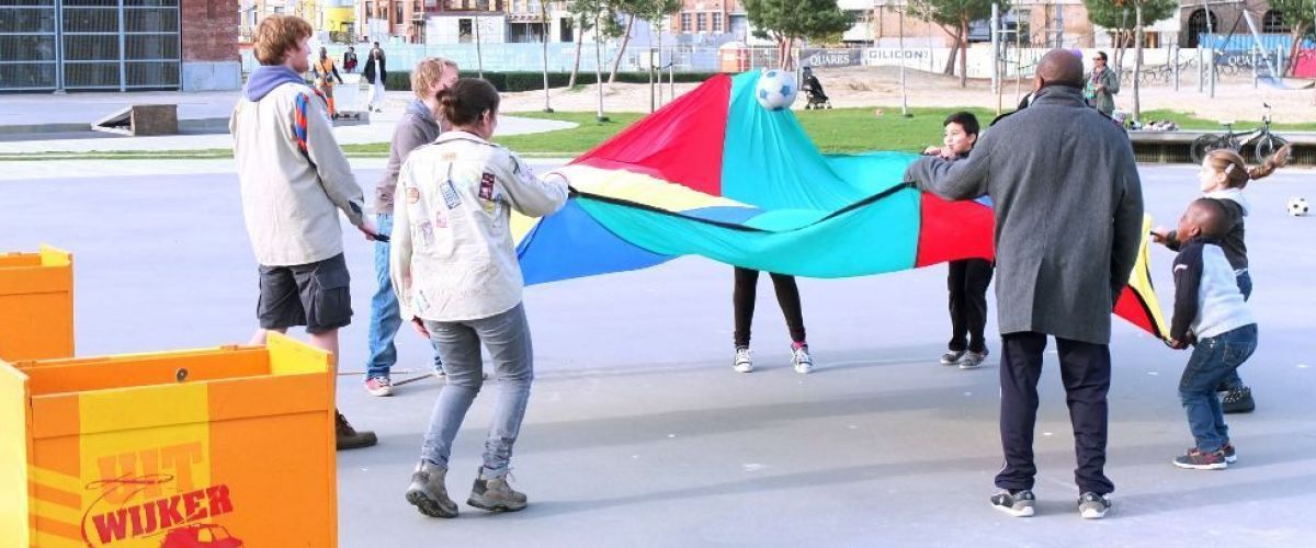 Leiding en kinderen uit de buurt spelen op een pleintje. Op de voorgrond staat een grote Uitwijker-koffer met spelmateriaal in.