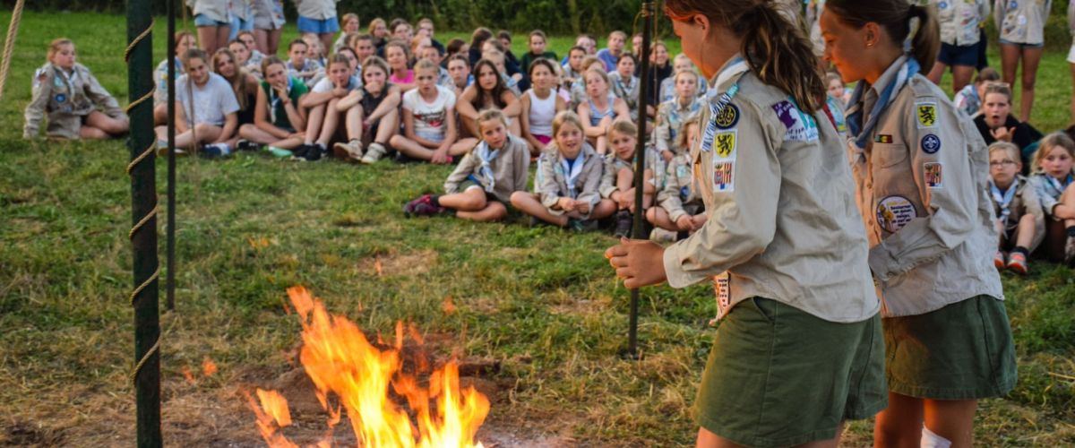 Twee gidsen staan aan het kampvuur. De rest van de scoutsgroep zit er rond.