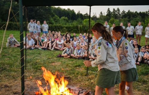 Twee gidsen staan aan het kampvuur. De rest van de scoutsgroep zit er rond.