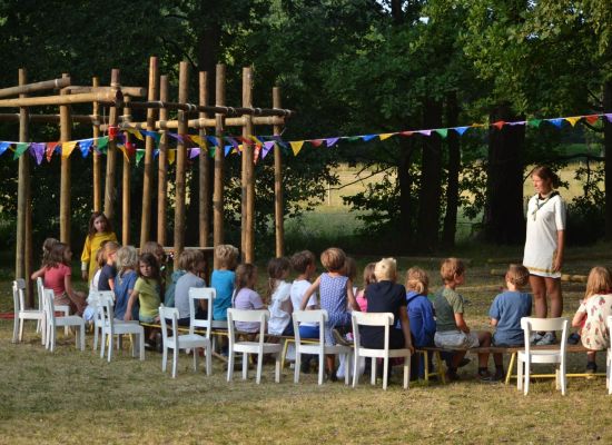 kapoenen op stoeltjes op een grasveld. Naast de kinderen staat een sjorconstructie versiert met vlaggetjes.