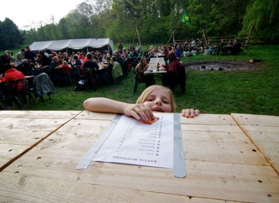 Op de achtergrond zitten mensen aan lange tafels, vooraan komt een kindje bestellen aan de toog, hoofd net boven de tafel.