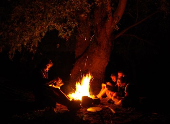 Scoutsleider leest voor aan drie leden rond het kampvuur