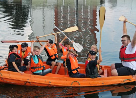 acht zeewelpen in een oranje bootje. Op de boeg zit een leider. Ze zwaaien met hun roeispanen.