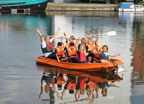 Kinderen met begeleider die met reddingsvesten in een raft zitten.