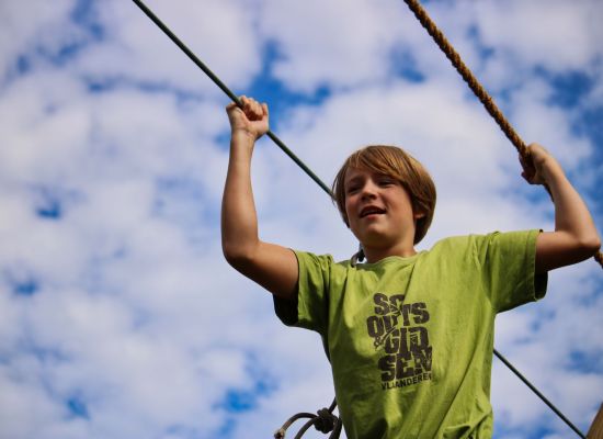 Een scout met groene scoutsshirt balancerend op een touwbrug