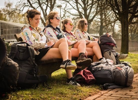 vier gidsen op een bank in het zonnetje, benen gekruist, rond hen rugzakken op de grond.