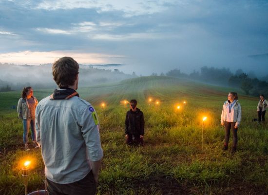 In een glooiend landschap bij zonsopgang zit een geblinddoekte scout op zijn knieën klaar voor zijn totem. Een leider spreekt hem toe.