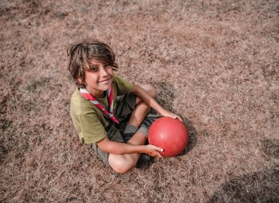 jongen met groene scoutst-shirt en korte groene broek zit in het gras met een rode bal.