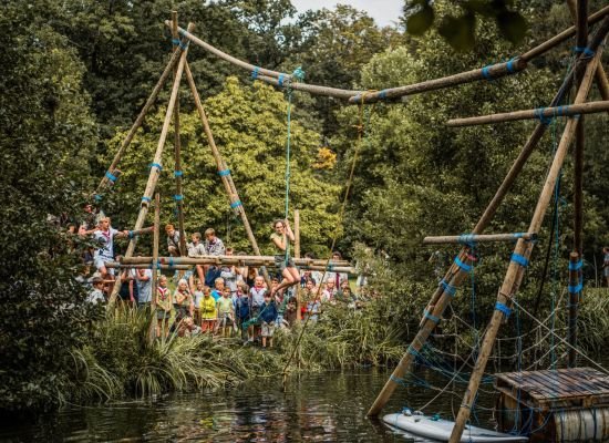 een grote sjorconstructie over een rivier met vanboven een touw waar iemand aan hangt die over de rivier zwiert.