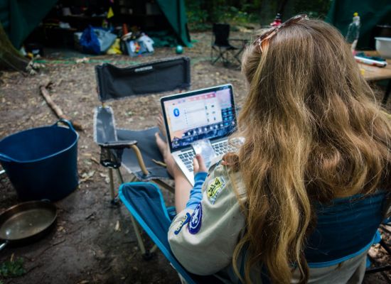 een leidster zit midden het bos voor een tent op haar laptop te werken