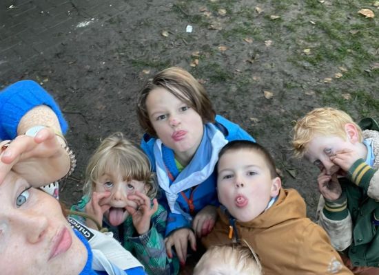 een leiding kijkt in de lens en doet de scoutsgroet. Kapoen kijken mee en trekken gekke bekken.