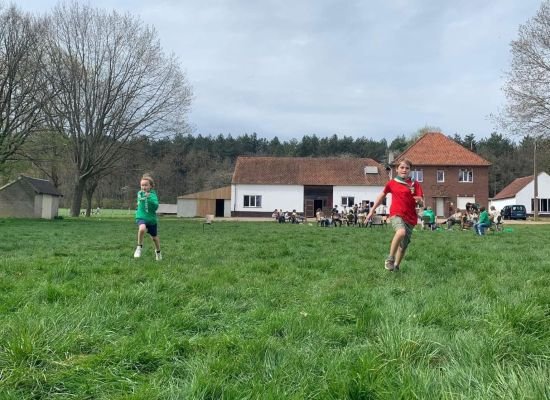 Leden lopen op een scoutsterrein, op de achtergrond zie je het lokaal