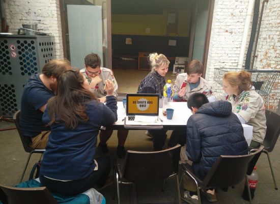 leiders en leidsters rond de tafel, in vergadering. Op tafel een opengeklapte laptop met op het scherm de grote KOS-quiz.