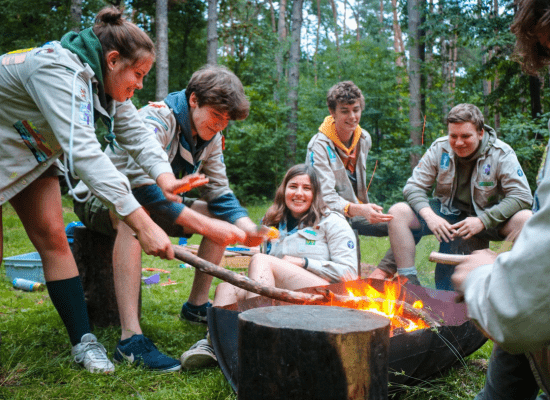 Een groepje leiding warmt zich ontspannen rond een vuurschaal. Een leidster houdt een tak in het vuur.